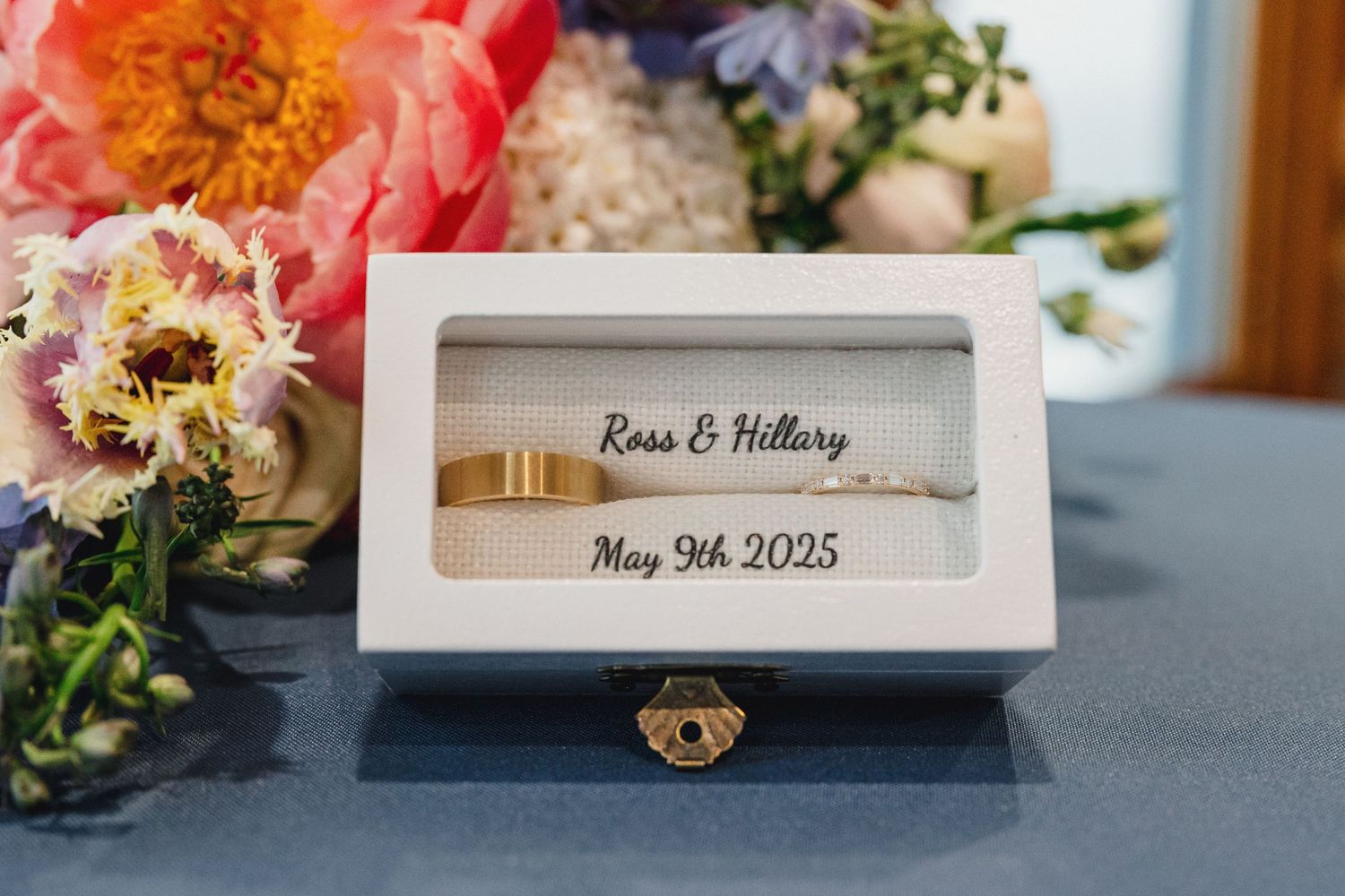 White wooden ring box with engraved names and date displayed next to colorful floral arrangements on blue surface.