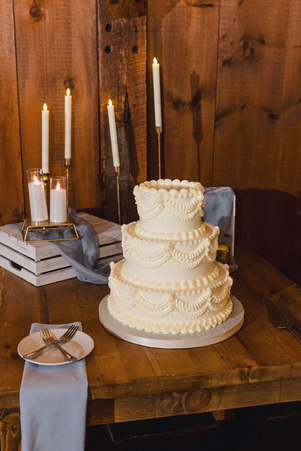 Three-tiered white wedding cakes with delicate piping displayed on tables in a rustic wooden barn setting.