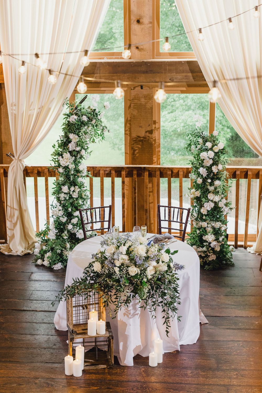 White draped curtains and floral arrangements decorate a rustic barn wedding venue with wooden floors.