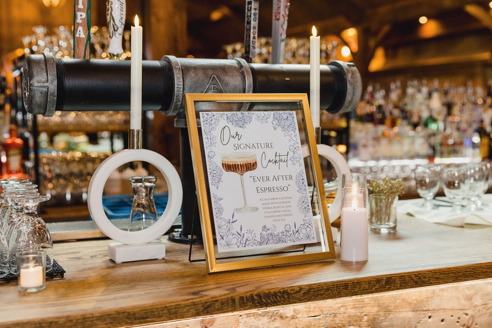 Bar setup with signature drink menu and glassware at rustic wedding reception.