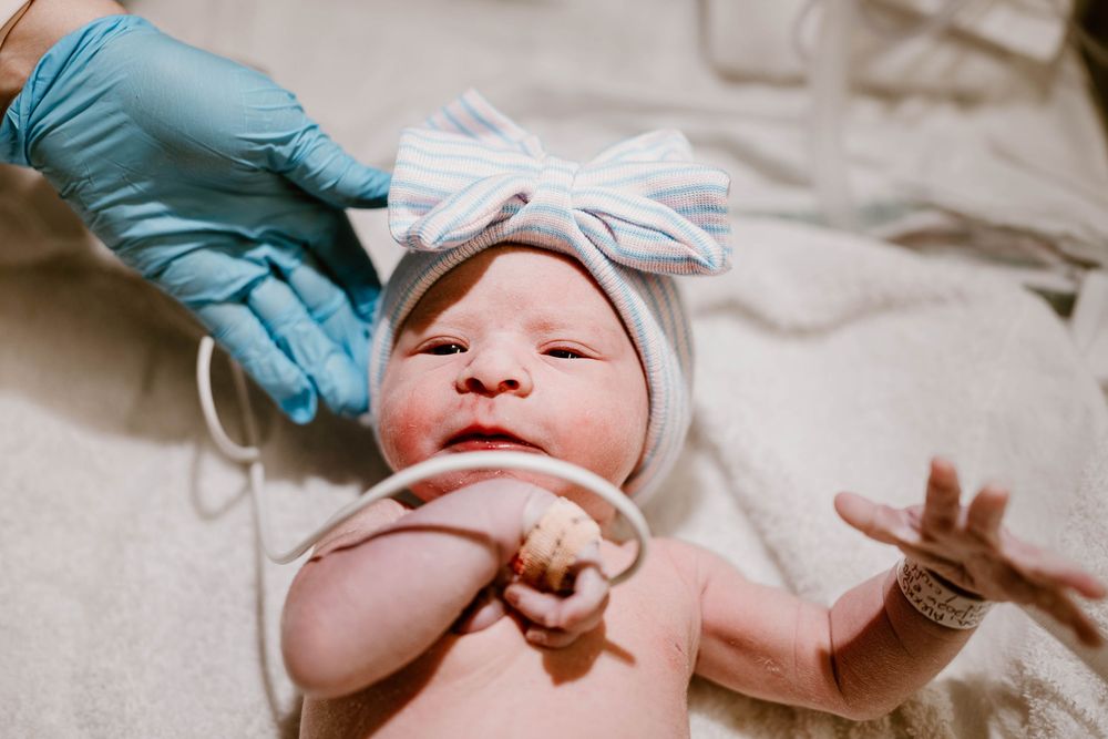Medical professionals in blue gloves examine a newborn baby wearing a white bow headband.
