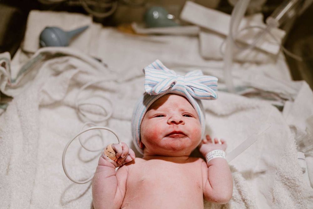 A newborn baby lies on soft white bedding wearing a light blue bow headband.