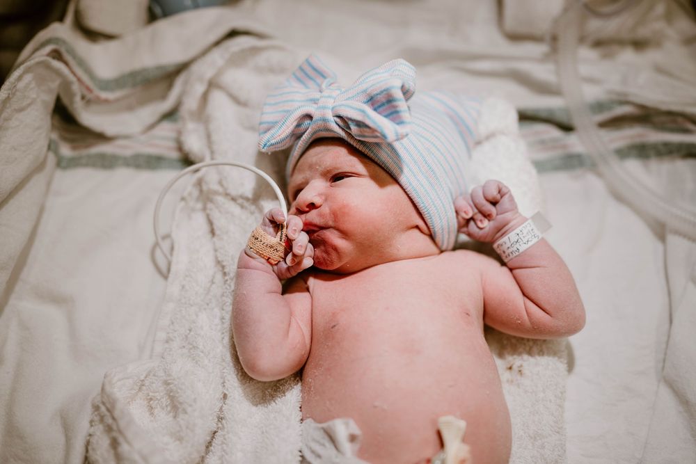Newborn baby wearing a light blue hat lies on beige floral bedding with arms raised.