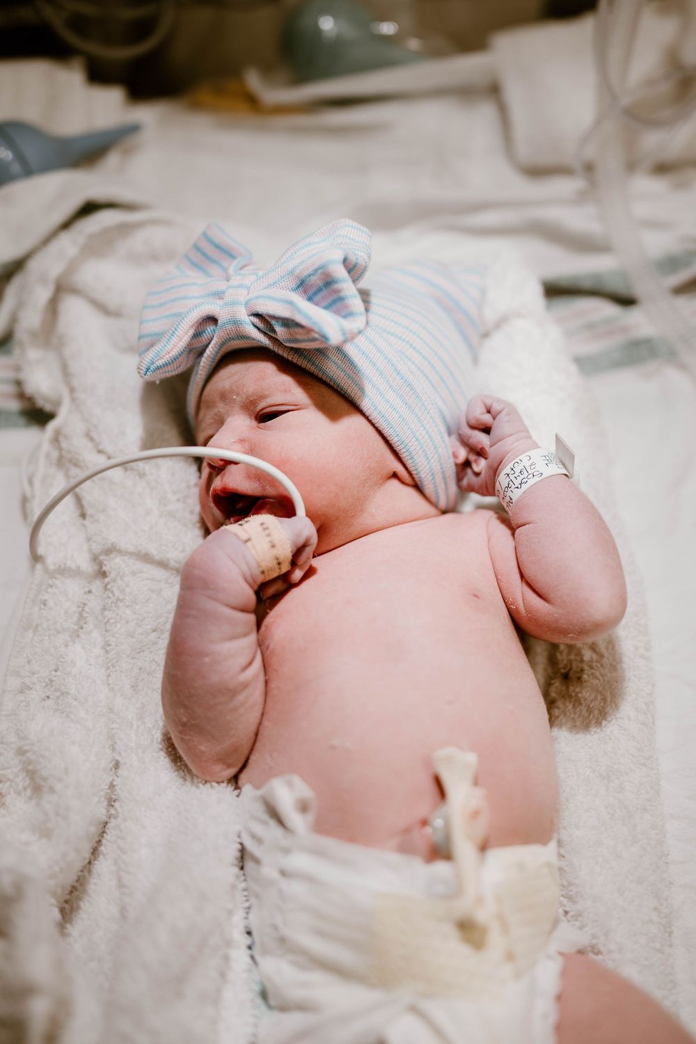 A newborn baby wearing a white hat lies on soft white blankets while yawning and stretching.