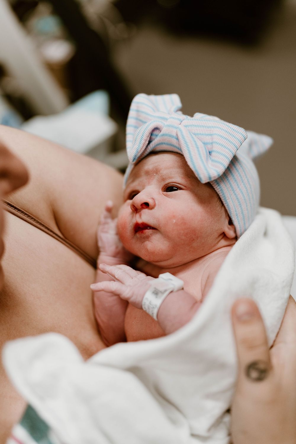 A newborn baby wearing a striped bow hat rests against someone's chest wrapped in a white blanket.