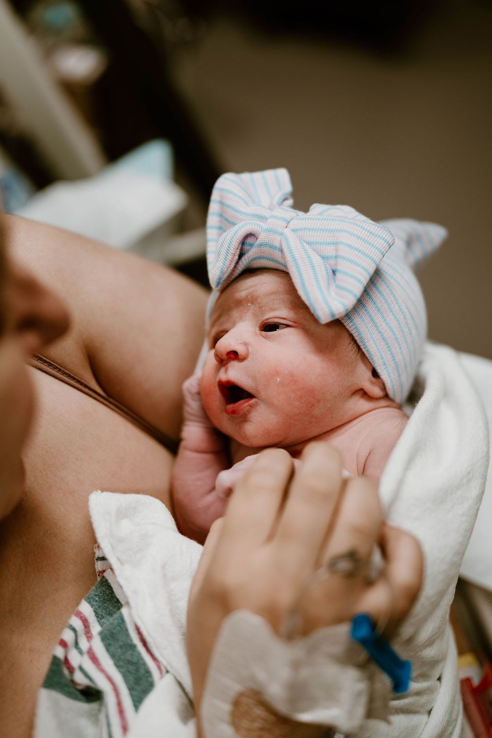 A newborn baby in a hospital hat yawns while being cradled in someone's arms.