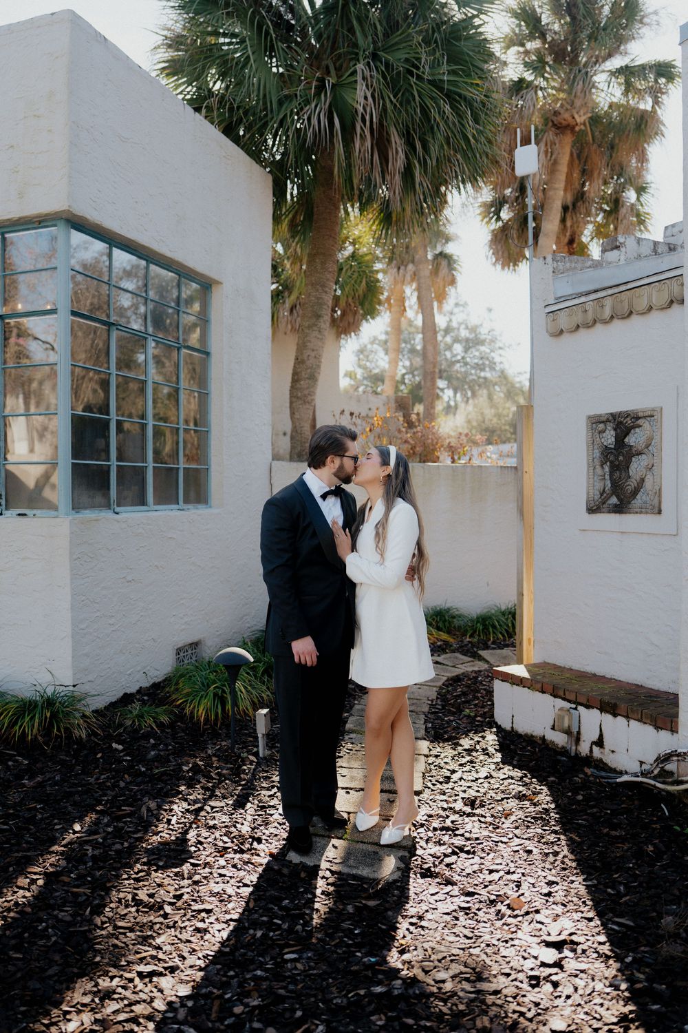 A couple in wedding attire share an intimate moment in a garden courtyard with palm trees and white walls.