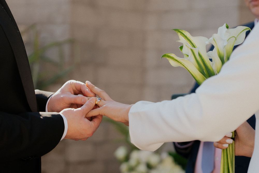 Close-up view of hands exchanging wedding rings during ceremony with white lily bouquet in background.