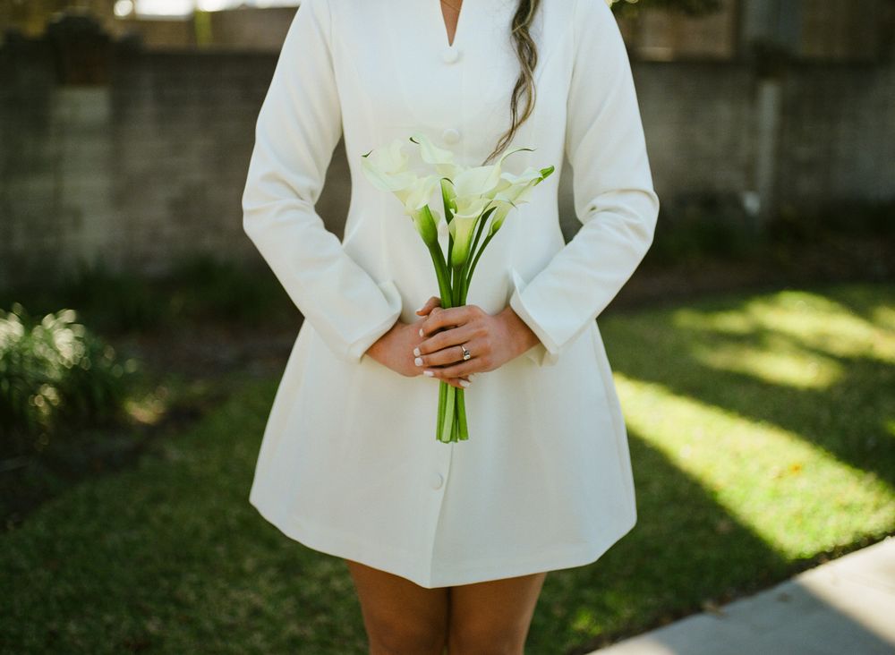 A person in a white blazer and shorts holds a small bouquet of white calla lilies in a sunny garden setting.