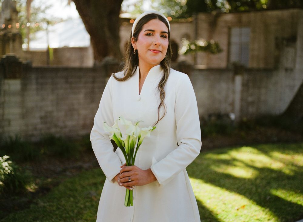 A bride in a white dress holds a simple bouquet of white calla lilies in a sunlit garden setting.