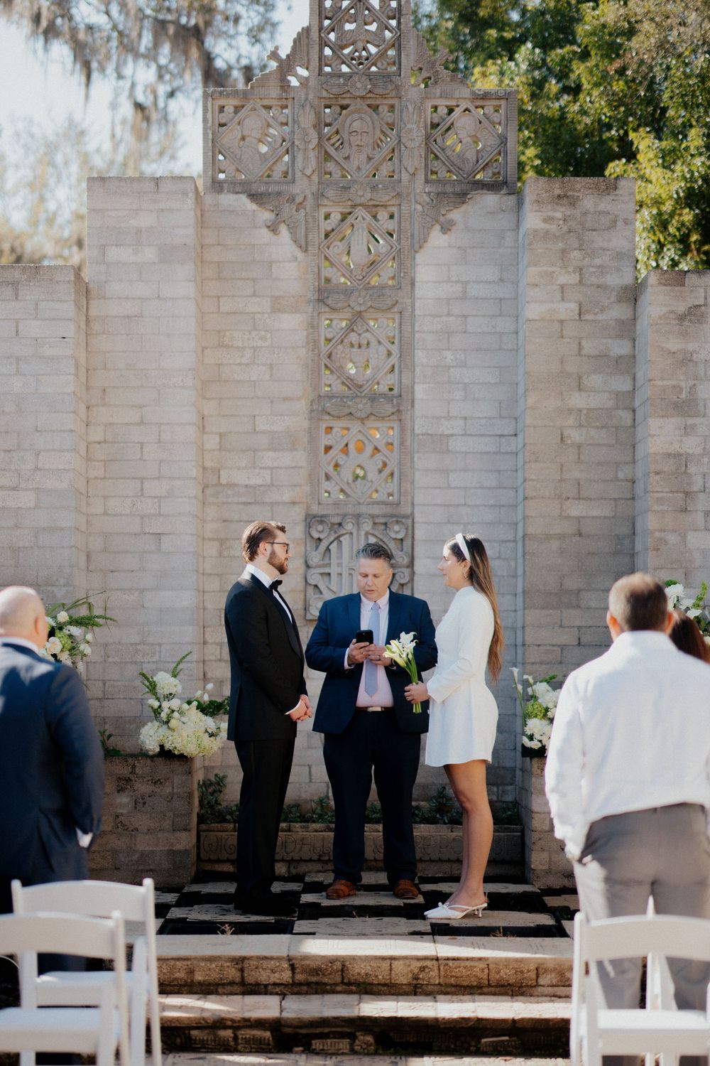 Small intimate outdoor wedding ceremony with white chairs and decorative stone wall backdrop.