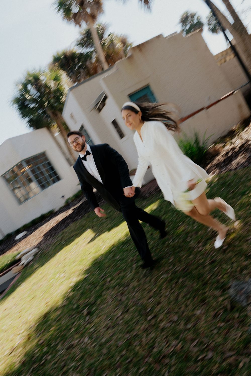 A couple runs playfully on a lawn in front of a white stucco house, with palm trees visible in the background.