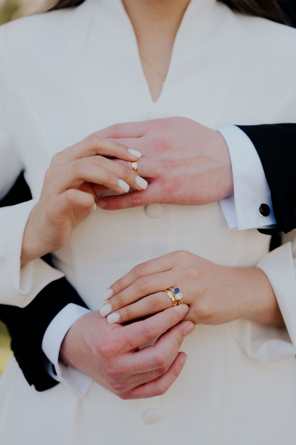 Close-up of hands wearing wedding rings against white and black formal attire.