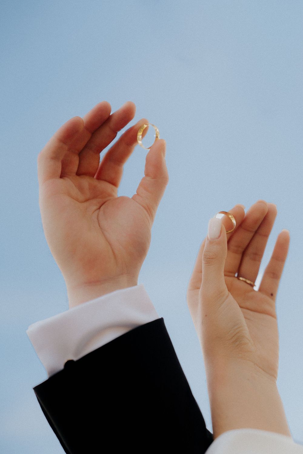 Close-up of hands wearing formal attire holding wedding rings against a blue sky background.