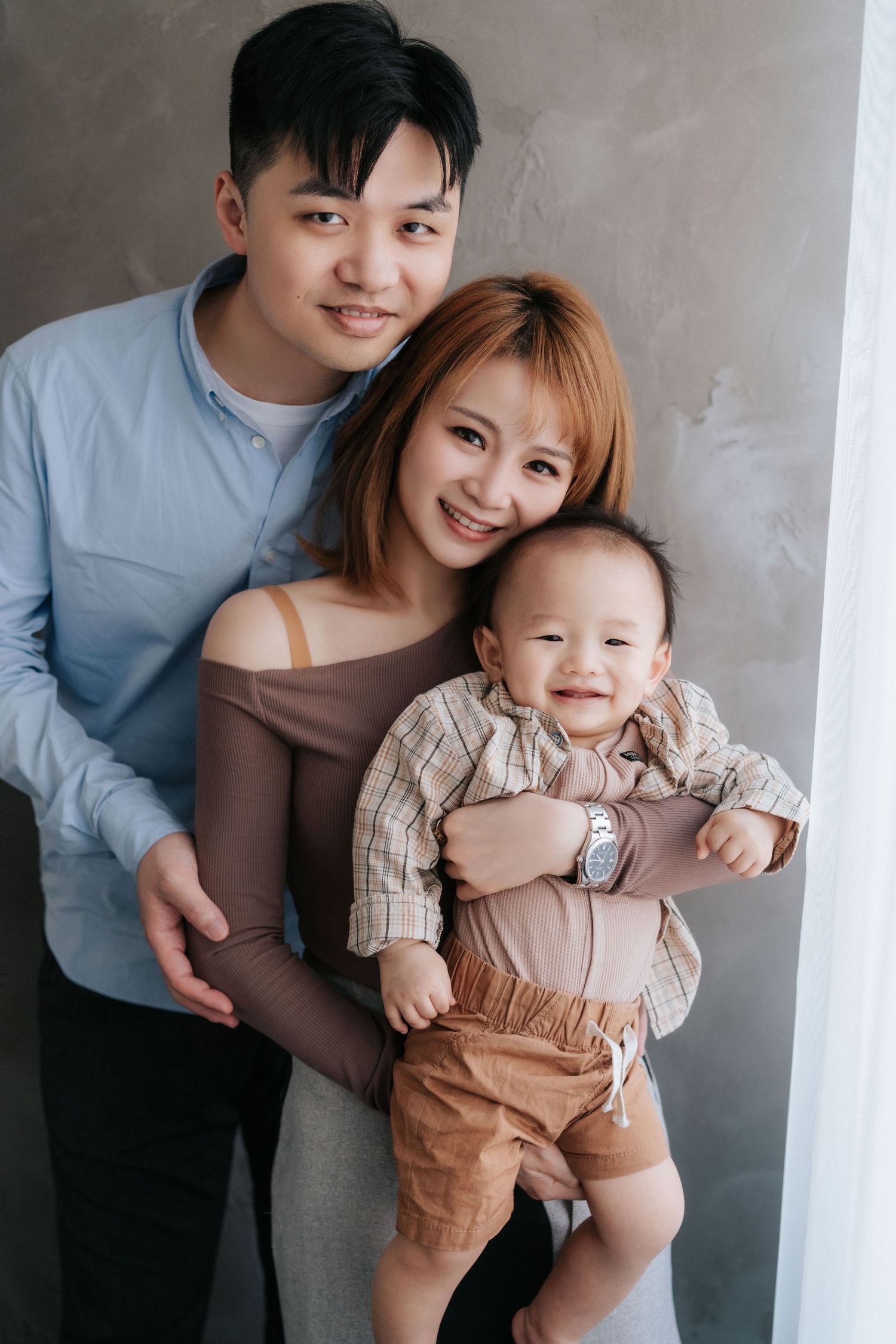 A family poses together against a gray wall in a series of candid lifestyle portraits in neutral earth tones.在一系列以中性大地色調為主的自然生活照中，一家人靠在灰色的牆壁