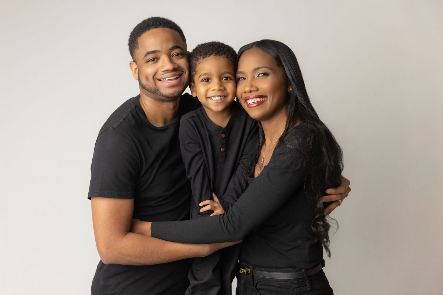 Three people wearing black outfits pose together for a family portrait against a light gray background.