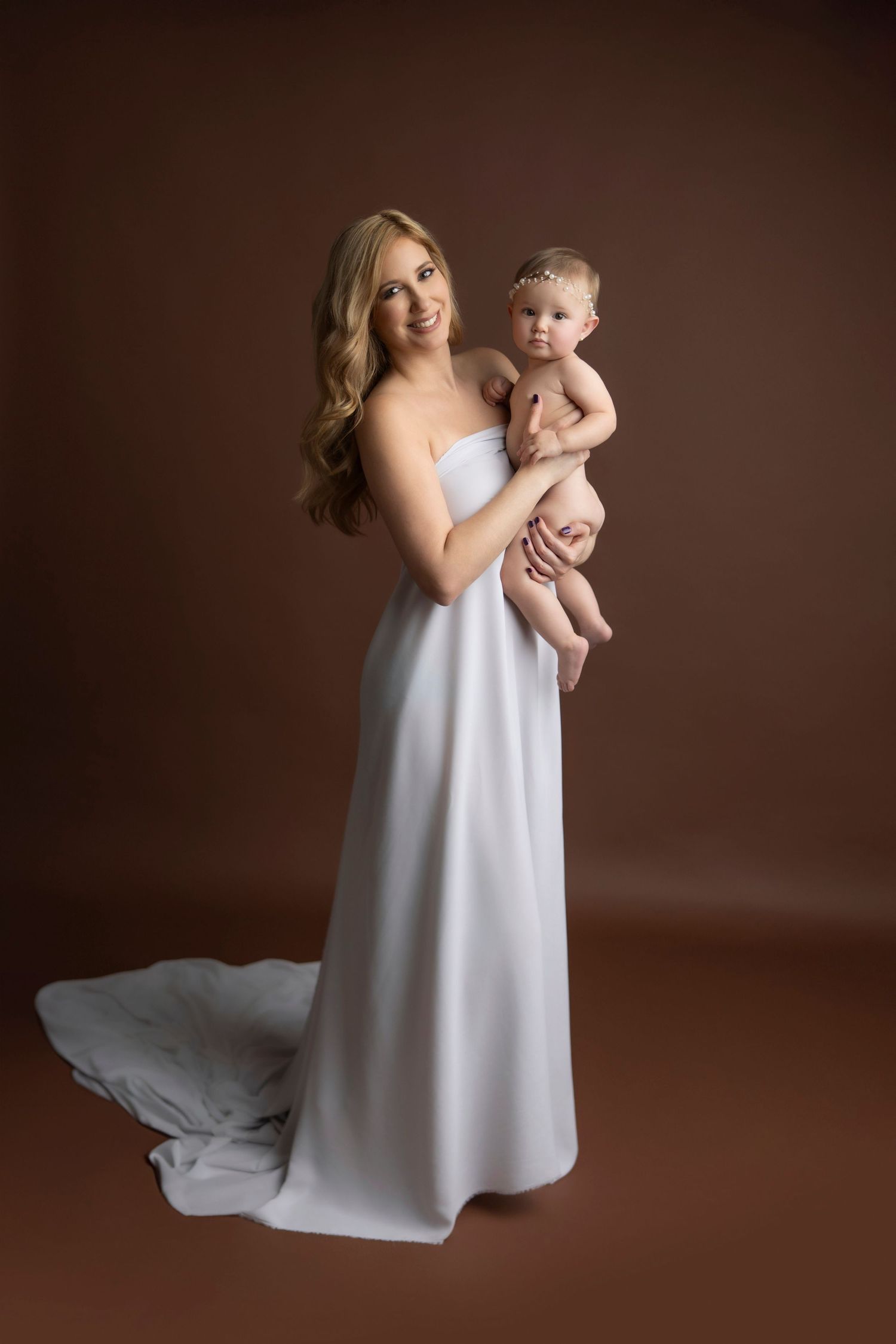 A parent in a flowing white dress holds a baby against a dark brown studio backdrop in a series of artistic portraits.