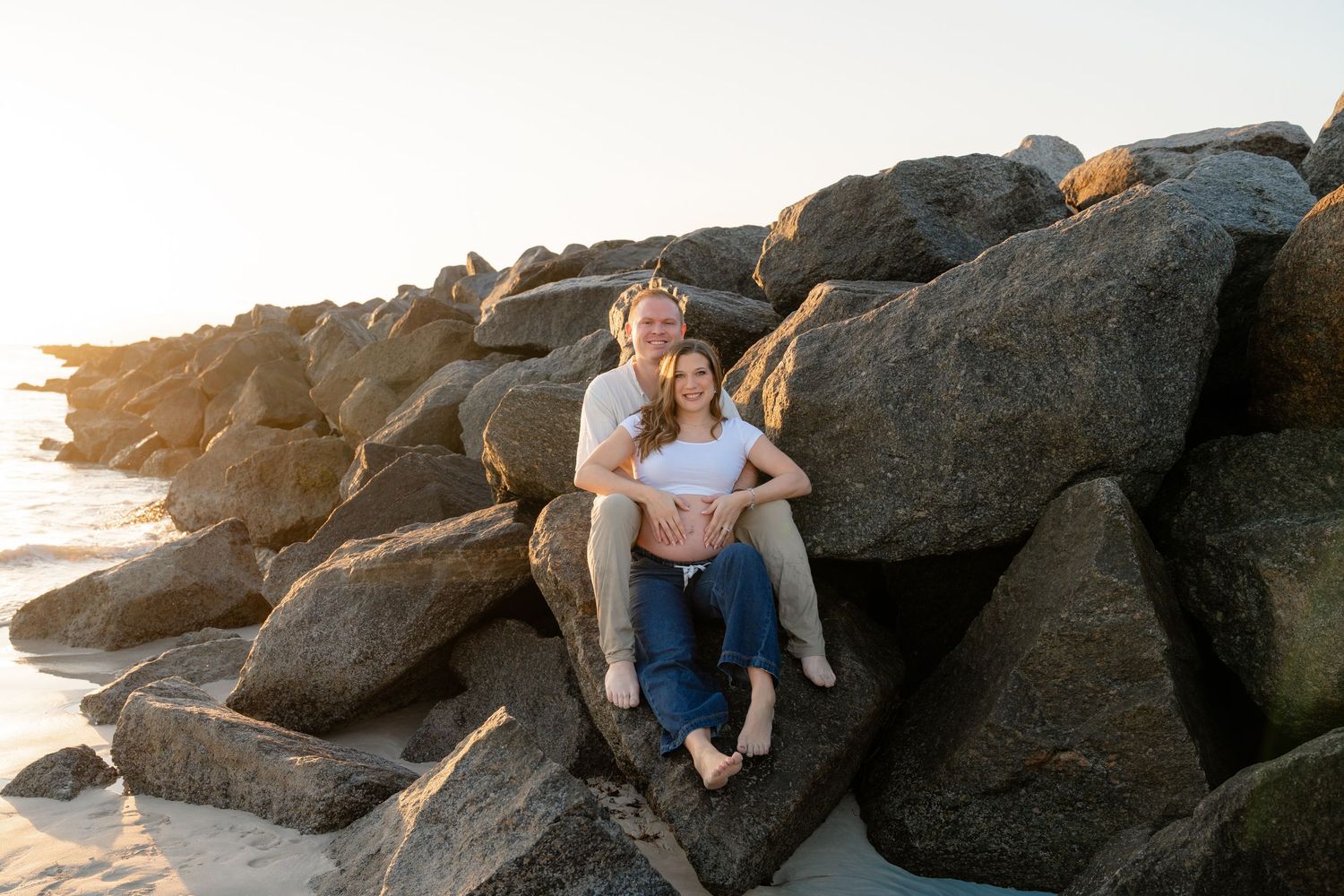 A couple sits together on coastal rocks during golden hour sunset at the beach.