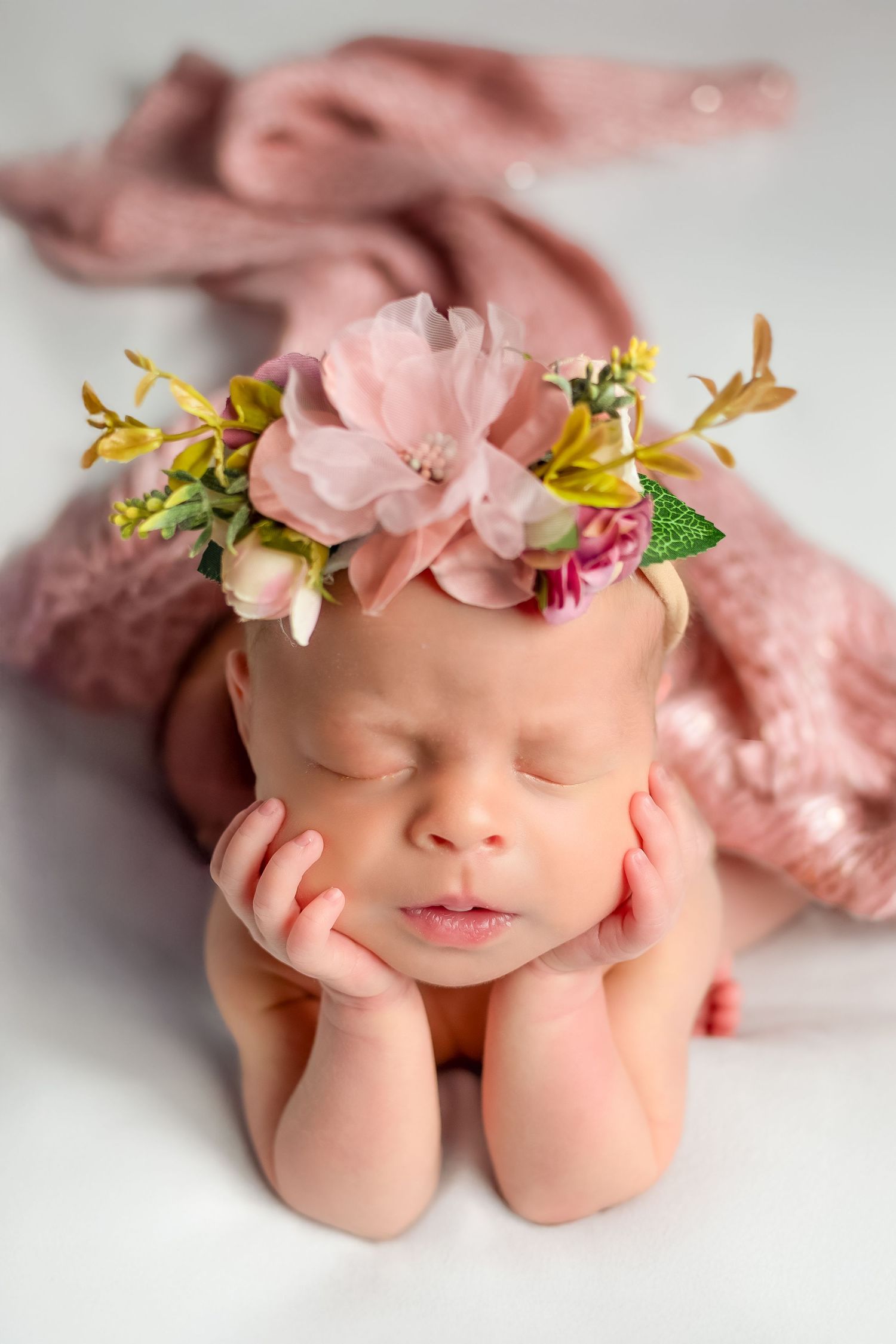 Newborn photography with delicate pink floral headpiece and soft fabric backdrop in gentle pastel tones.