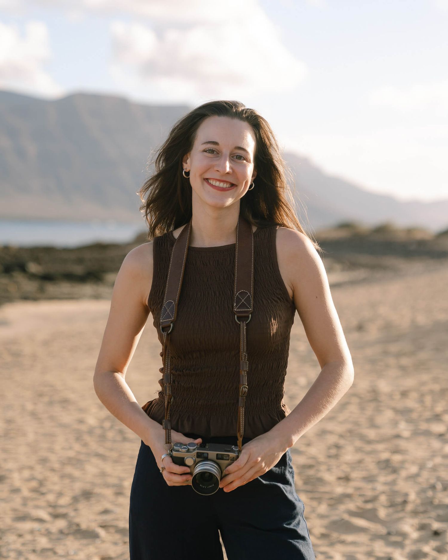 A photographer with a vintage film camera stands smiling on a sandy beach with mountains in the background.