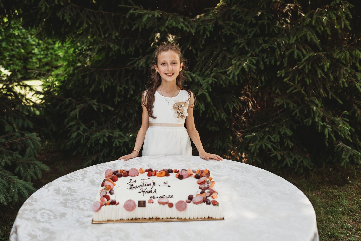 A white celebration cake sits on a table outdoors against a backdrop of green foliage.