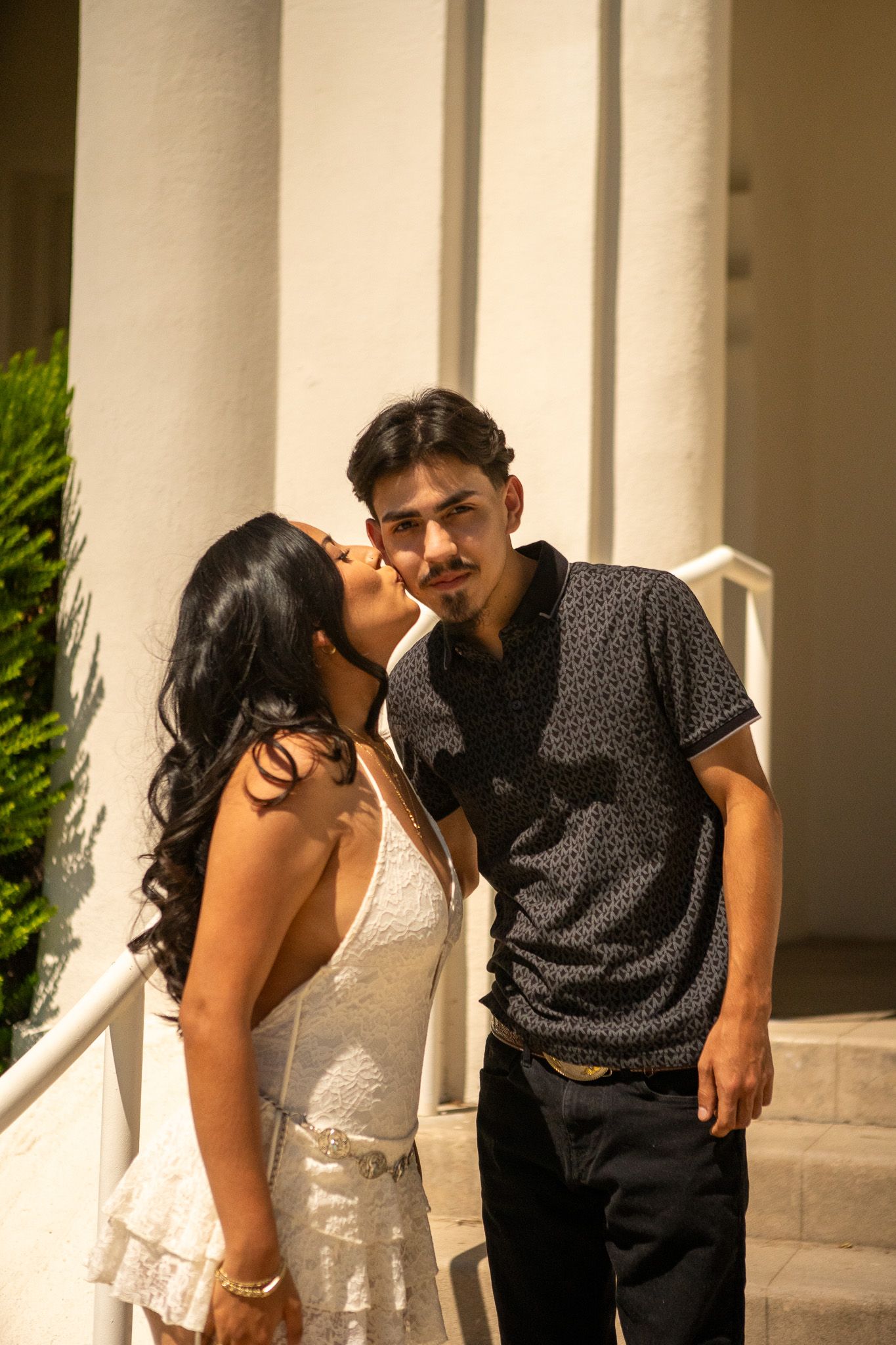 A couple shares an intimate moment while posing together against white columns in an outdoor setting.