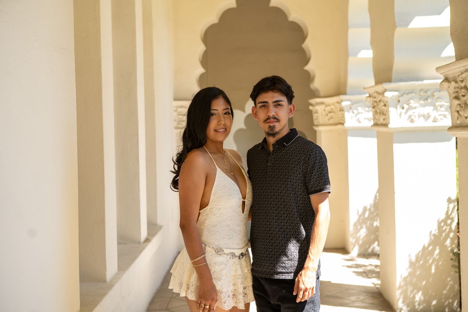 A couple in wedding attire poses together in a bright white hallway with sunlight streaming through arched windows.