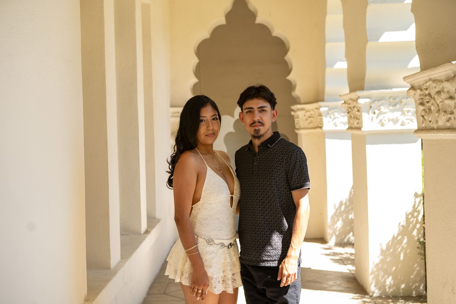 A couple stands in a bright sunlit hallway with white columns and dappled light creating a romantic atmosphere.