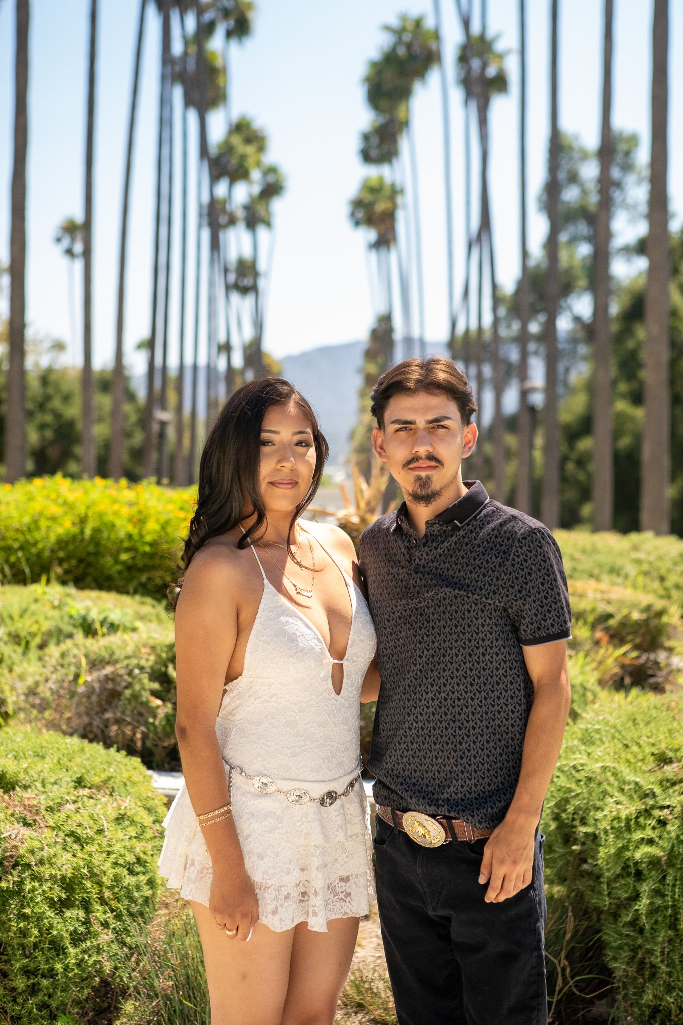A couple poses together in a sunny outdoor setting with palm trees and lush greenery in the background.