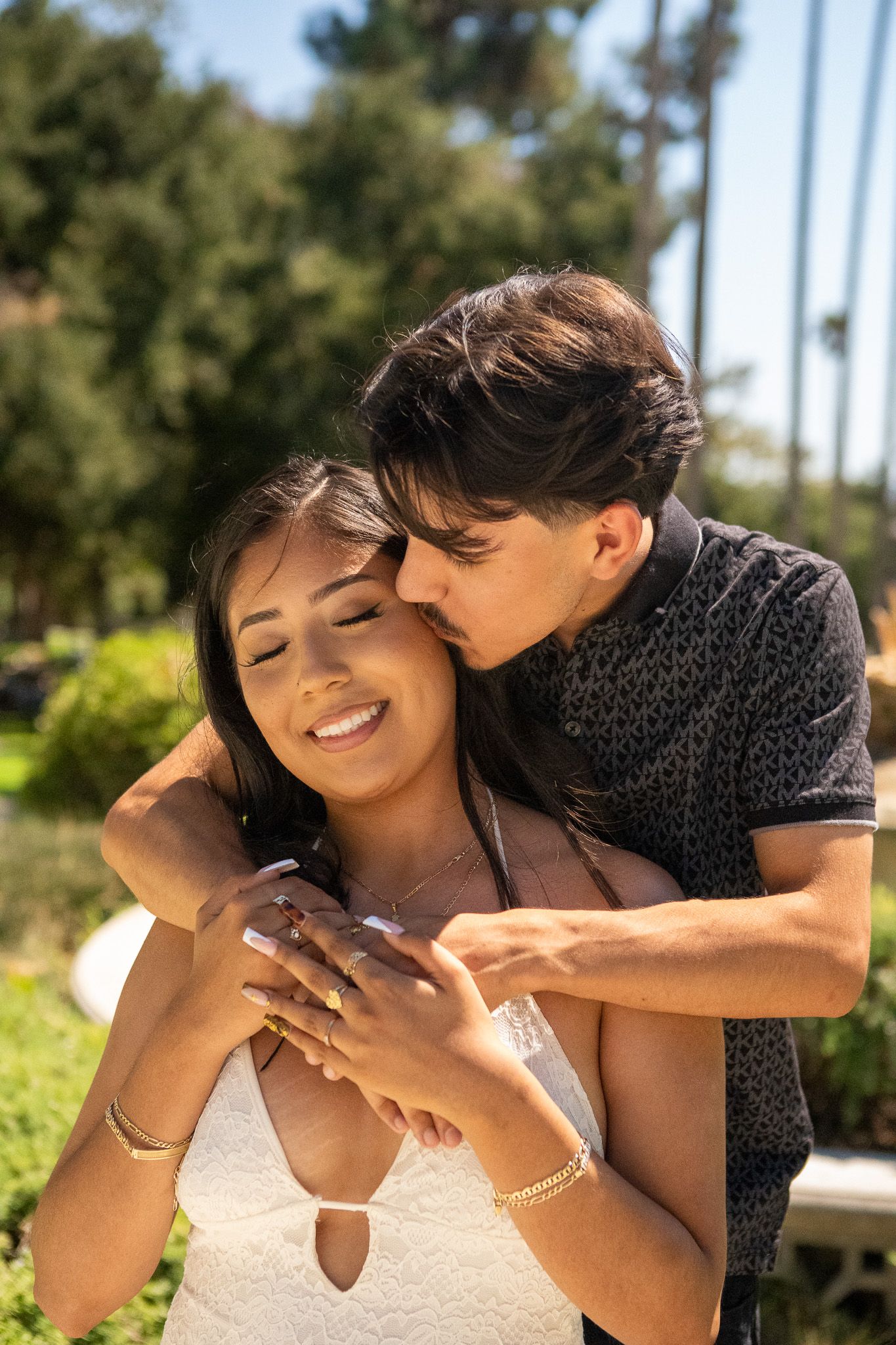 A romantic couple shares an intimate moment outdoors on a sunny day with trees in the background.