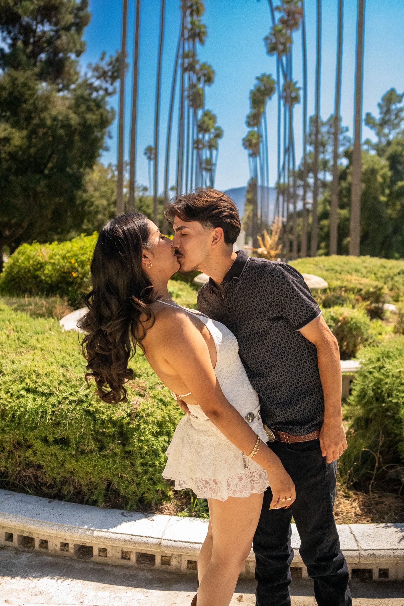 A romantic couple shares a playful dip kiss pose among tall palm trees in a sunny garden setting.