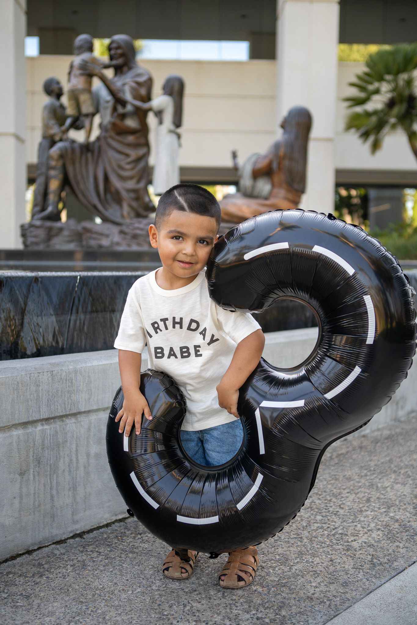 A young child in a birthday t-shirt poses with a large black inflatable tube outdoors near a stone wall.