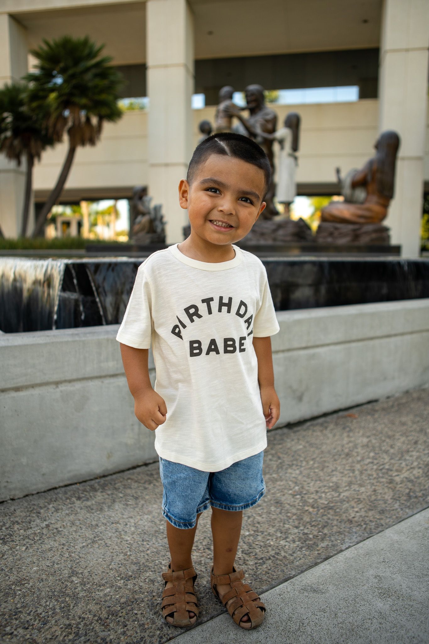 A small child wearing a white t-shirt and denim shorts stands on a concrete surface outdoors.