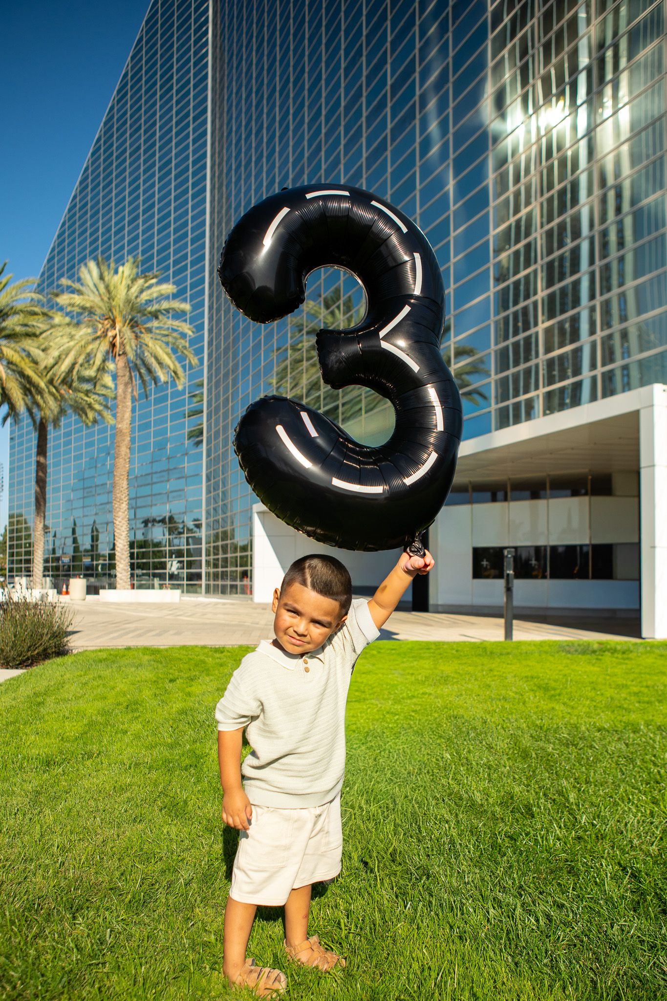 A child holds up a large black number 3 balloon in front of a modern glass office building with palm trees.