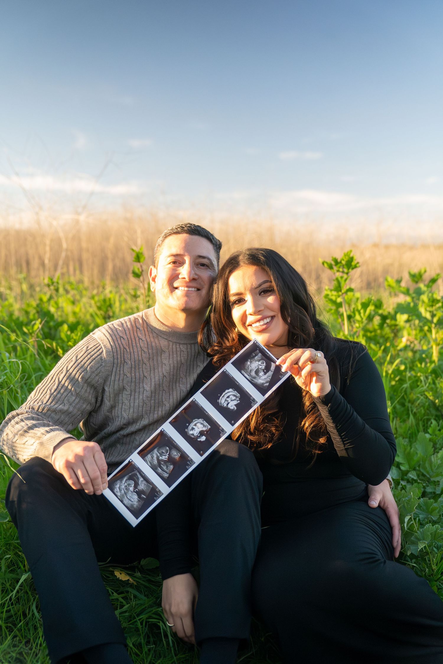 A couple laying in a grassy field sharing ultrasound photos during a pregnancy announcement photoshoot.