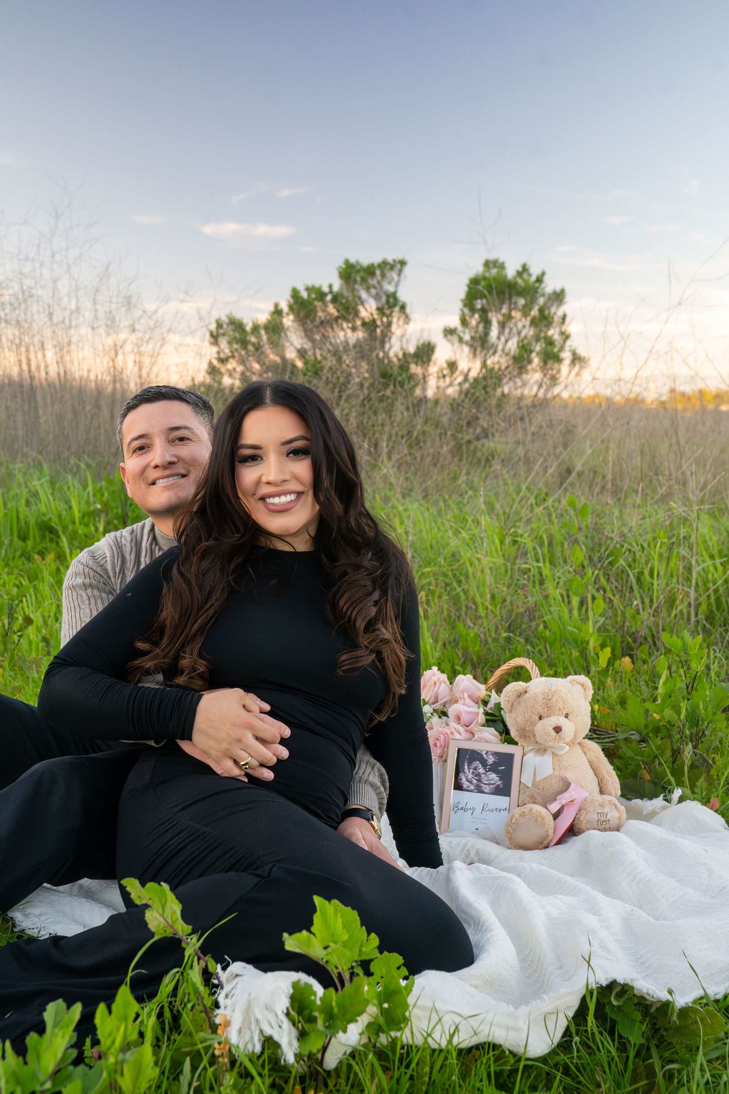 A couple sits on a white blanket in a grassy field at sunset with a teddy bear and ultrasound photo beside them.