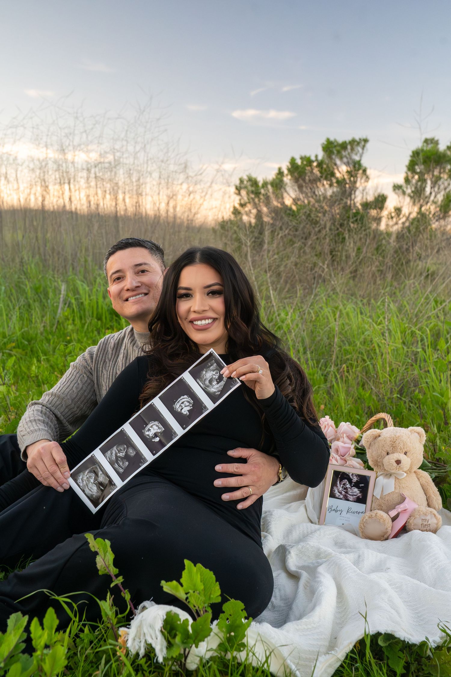 Couple sitting on white blanket in grassy field holding ultrasound photos and teddy bear during pregnancy announcement photoshoot.
