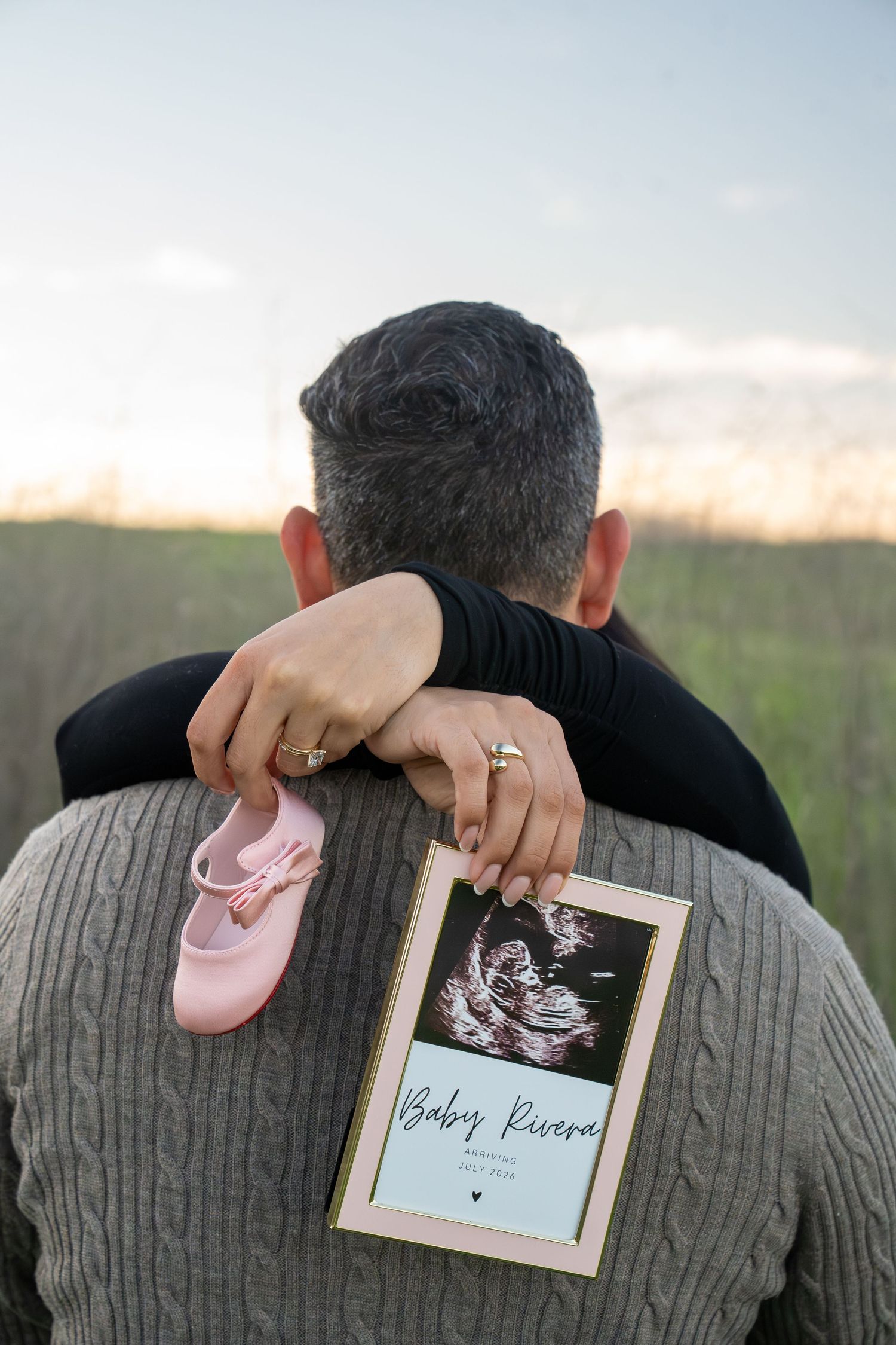 Pink baby shoes and ultrasound photos displayed on person's back during outdoor pregnancy announcement session.
