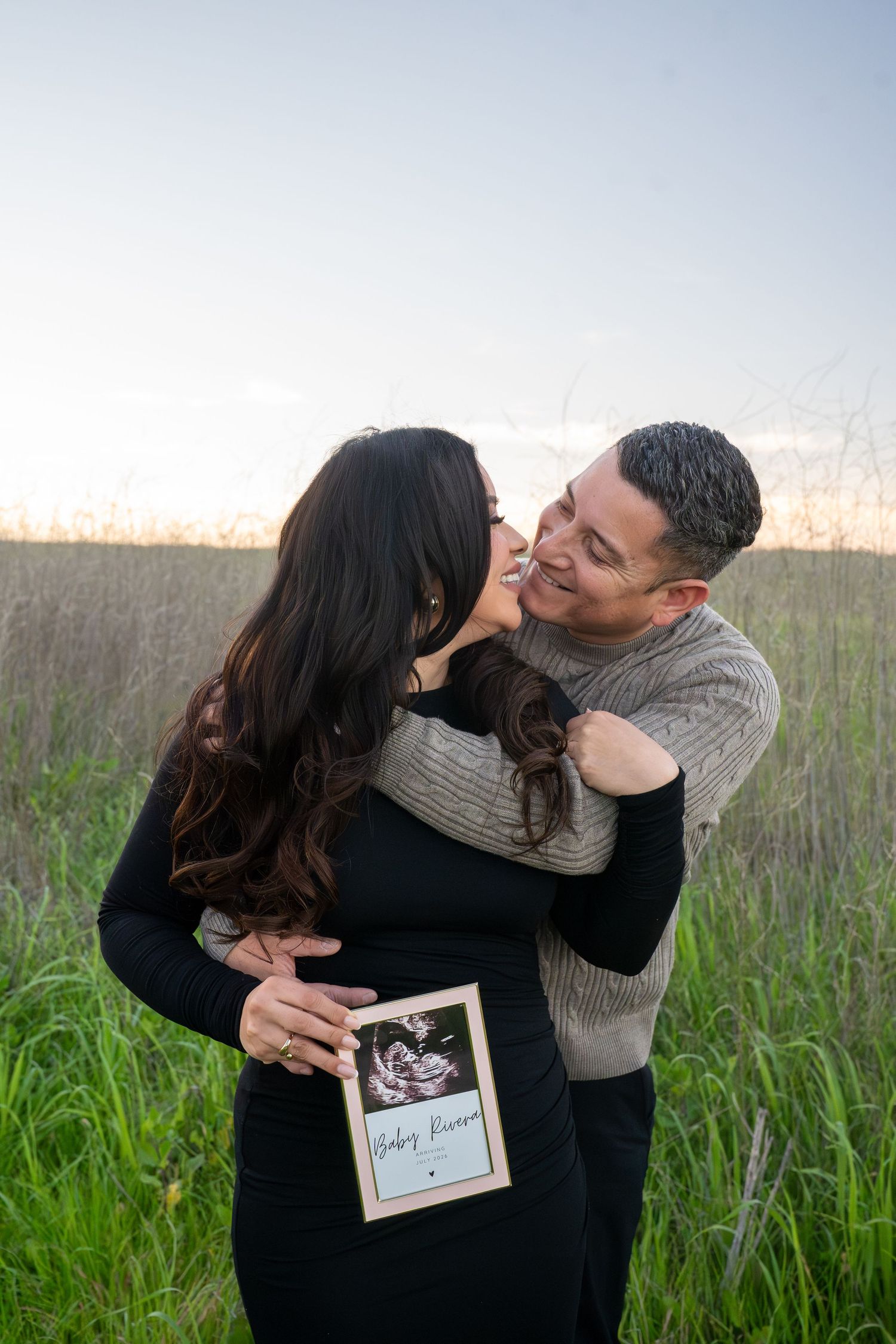 A loving couple embraces in a field while holding ultrasound photos during their pregnancy announcement session.