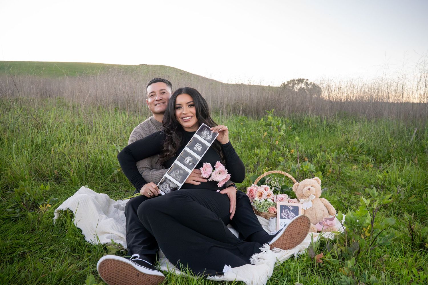 Couple celebrating pregnancy announcement with ultrasound photos and teddy bear in outdoor field setting.