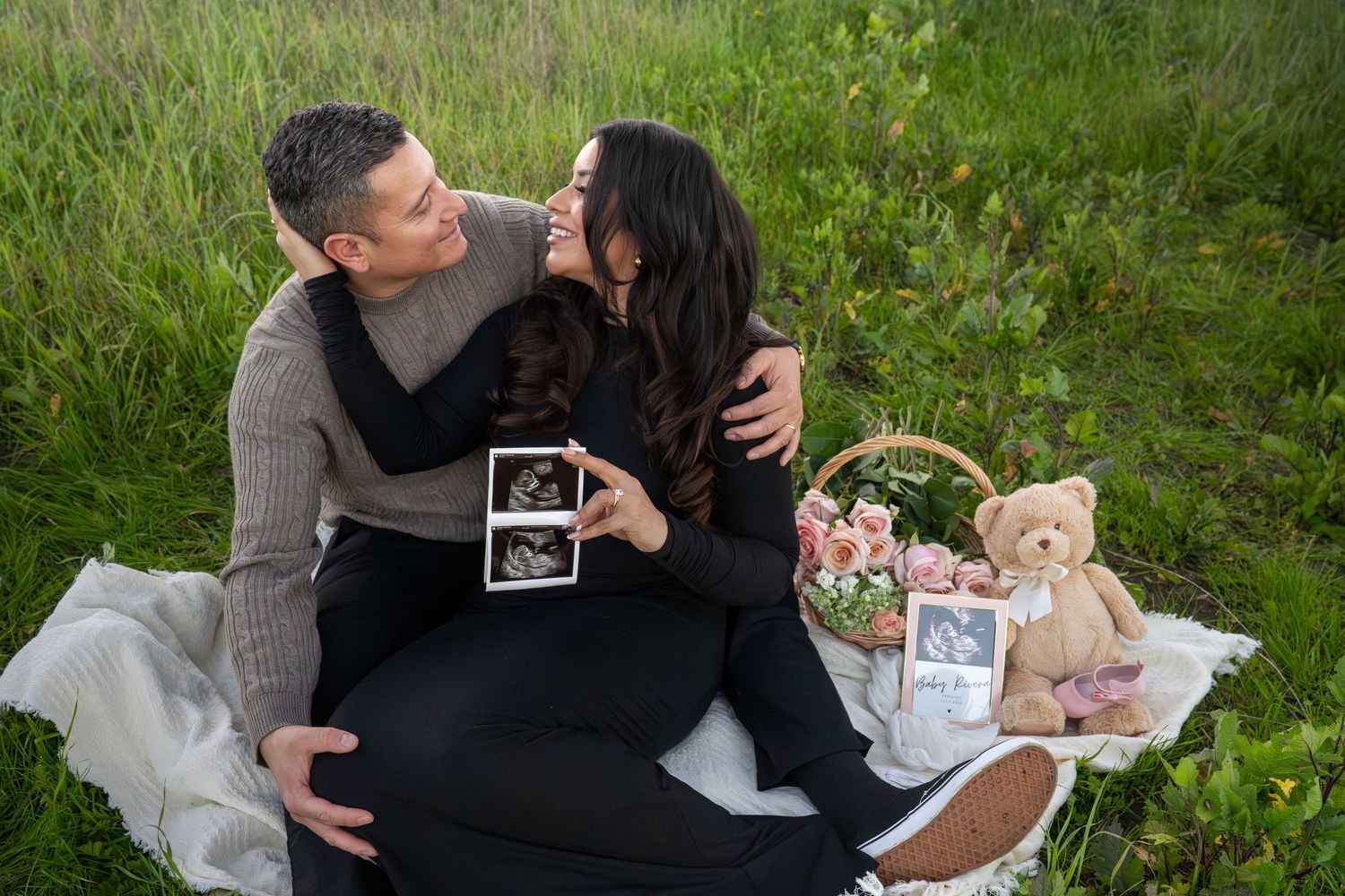 A couple shares a romantic moment during a picnic in a lush green meadow with a teddy bear and ultrasound photos nearby.