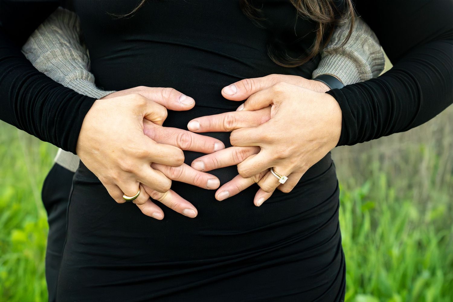 A person wearing black clothing makes heart shapes with their hands against a green grassy background.