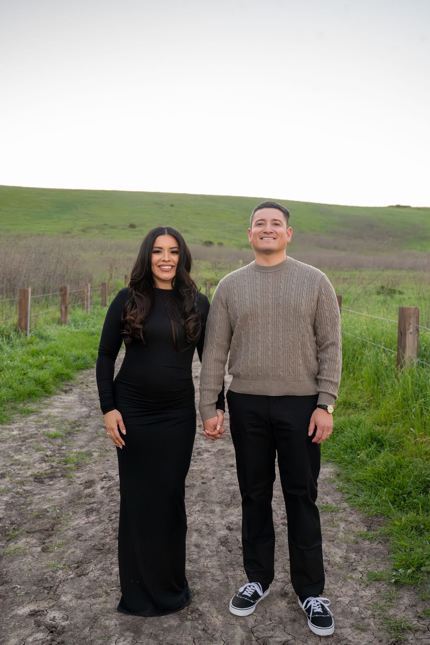 Couple in coordinated black outfits standing together on a grassy hillside path during an outdoor photo session.