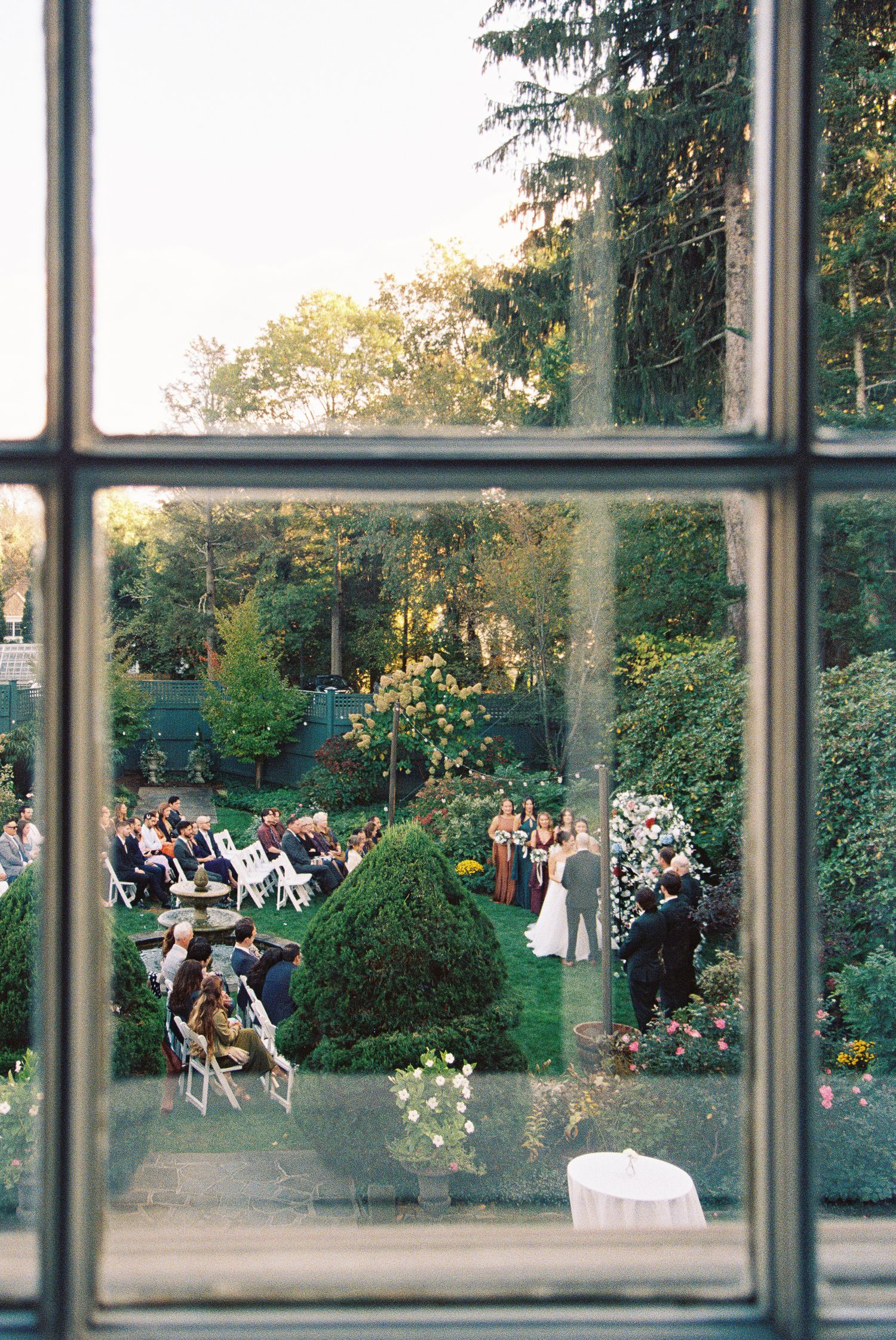 View through window panes of an outdoor garden wedding ceremony with guests seated on white chairs among lush greenery.