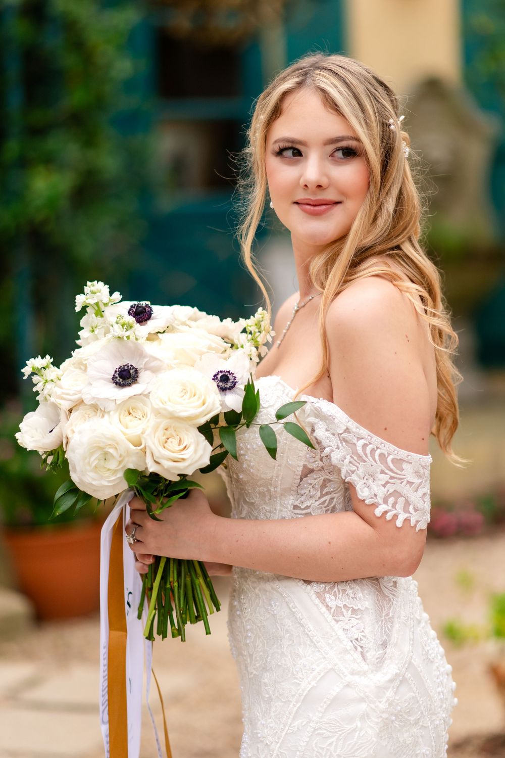 A bride in a white lace wedding dress holds a bouquet of white roses and anemones against a garden backdrop.
