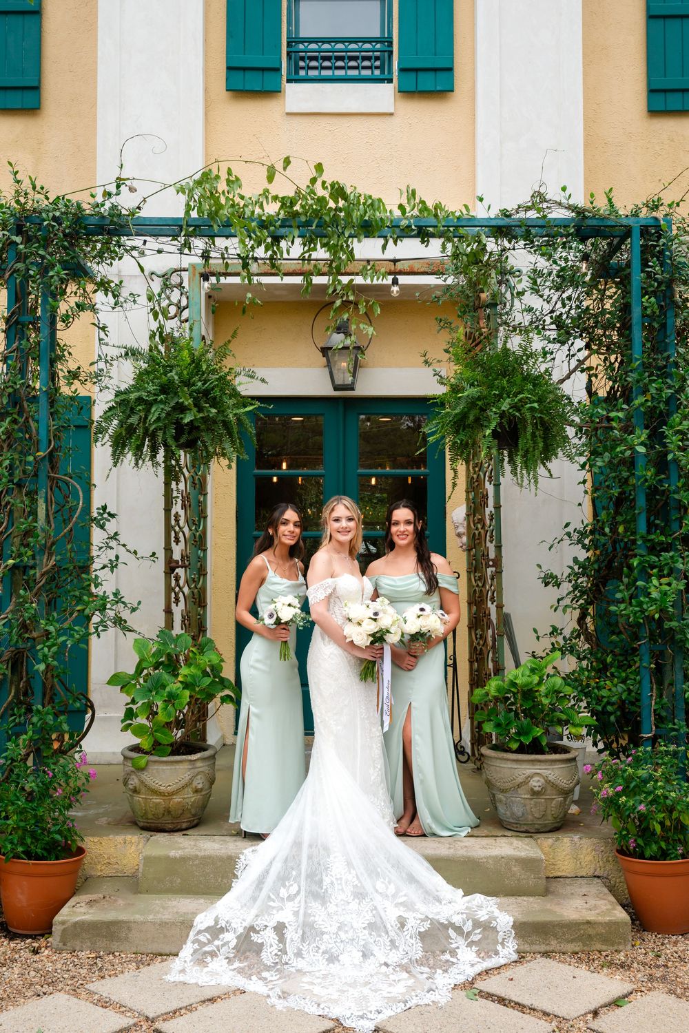 Elegant wedding party poses in front of cream colored building with green shutters and potted plants.