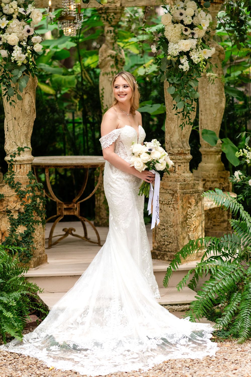 A stunning bridal portrait in a garden setting featuring a fitted off-shoulder lace gown with a dramatic cathedral train.