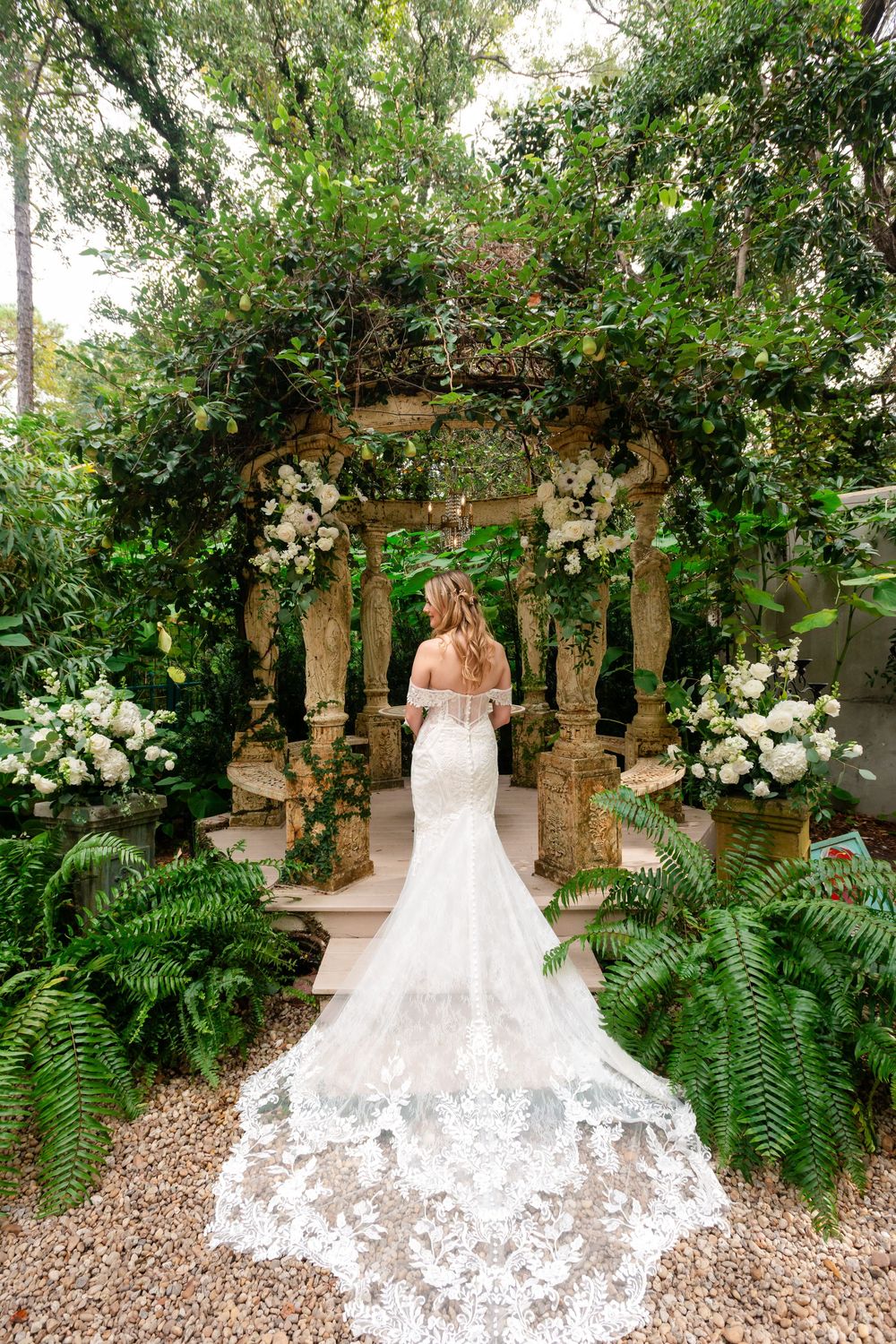 Wedding gown with intricate lace train displayed in lush garden setting with stone pillars and greenery.