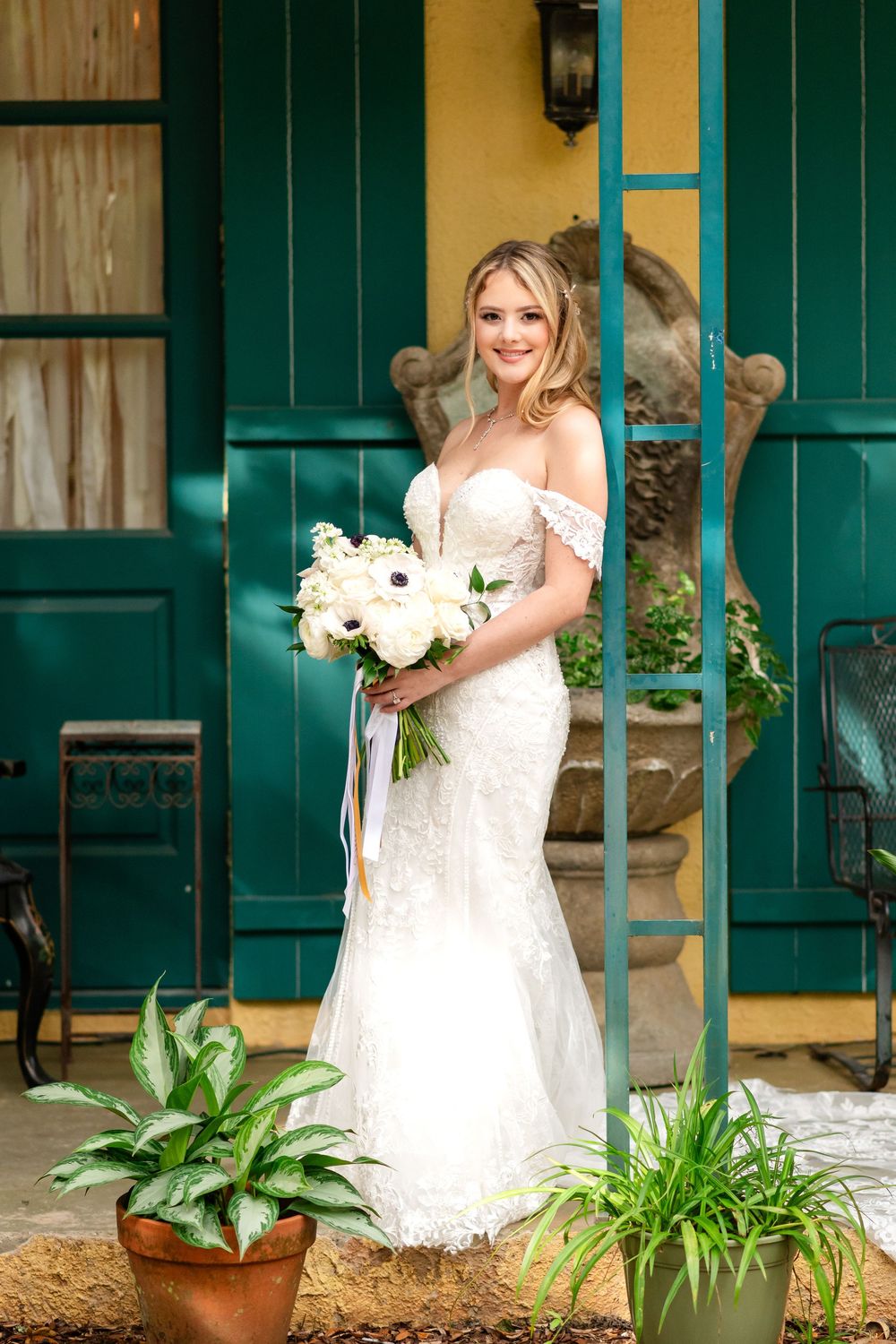 A bride in a white lace dress poses with a bouquet of white flowers on a porch with green shutters and potted plants.