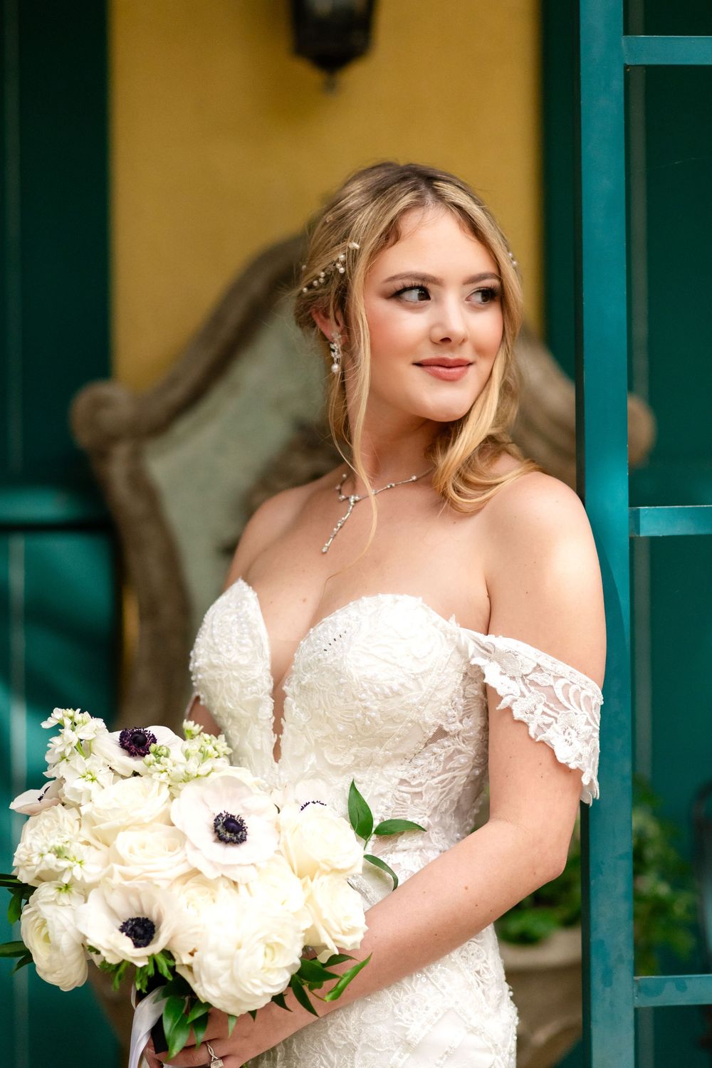 A bride in a white off-shoulder lace wedding dress holds a bouquet of white and purple anemone flowers.